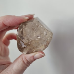May include: A faceted, translucent smoky quartz crystal. The crystal is held in a hand, showcasing its natural, light brown color and geometric shape. The crystal's surface reflects light, highlighting its clarity and internal structures.