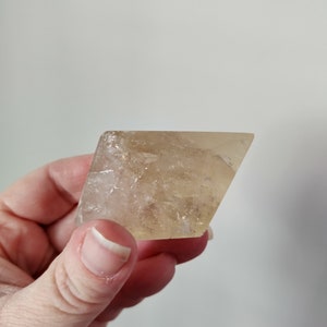 May include: A translucent, amber-colored crystal with a geometric shape. The crystal is held in a hand, showcasing its natural, raw texture and subtle variations in color. The crystal's edges are slightly irregular, adding to its unique appearance.