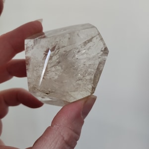 May include: A clear quartz crystal with a faceted, geometric shape. The crystal is held in a hand, showcasing its translucent quality and internal inclusions. The crystal's edges are polished, reflecting light.