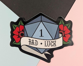 May include: A sticker featuring a blue, multi-sided die with the number 1. The die is surrounded by red roses and a banner that reads "BAD • LUCK". The sticker has a black border.