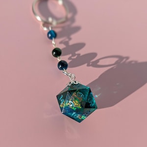Puede incluir: Un llavero de dados D20 turquesa y dorado con un anillo plateado. El dado está unido a una cadena de cuentas azules y verdes. El fondo es de color rosa liso.