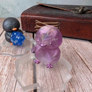 Puede incluir: Dos figuras de pingüinos, una negra y otra morada, están sobre plataformas transparentes y brillantes. El pingüino negro sostiene un dado azul de múltiples caras. El pingüino morado tiene un aspecto translúcido e iridiscente. Un libro encuadernado en cuero marrón está en el fondo.