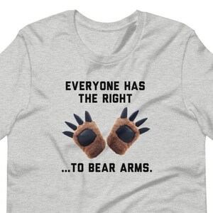 Può includere: T-shirt grigio chiaro con la scritta "EVERYONE HAS THE RIGHT...TO BEAR ARMS." La grafica presenta due guanti a forma di zampe di orso marroni. La maglietta è a girocollo e maniche corte.