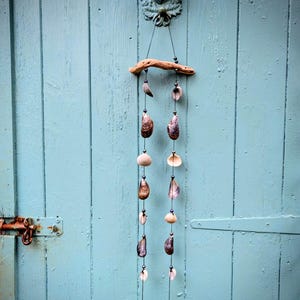 May include: A hanging mobile featuring a cast metal deer head hook at the top, from which two strings of dark beads, assorted natural seashells, and small pieces of driftwood are suspended. The mobile is composed of organic materials and metal.