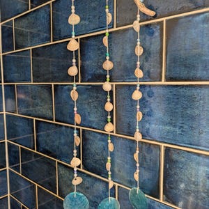 May include: A hanging mobile featuring a piece of driftwood at the top with three vertical strings attached. Each string is adorned with small white and green beads and various natural seashells, ending with a large, flat, circular blue shell.
