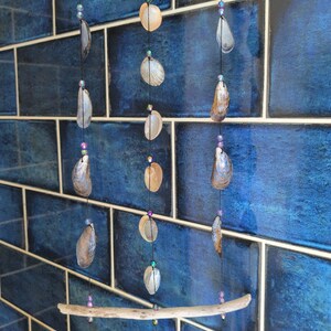 Puede incluir: Un móvil colgante que presenta dos piezas horizontales de madera flotante clara conectadas por cuerdas negras. Las cuerdas están adornadas con varias conchas marinas y pequeñas cuentas iridiscentes moradas y verdes en filas.