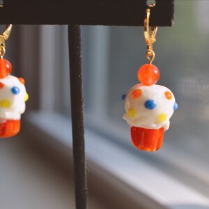 May include: A pair of whimsical cupcake earrings. Each earring features a white frosted cupcake with colorful polka dots, a red cupcake liner, and a small orange bead. The earrings have gold-colored leverback closures.