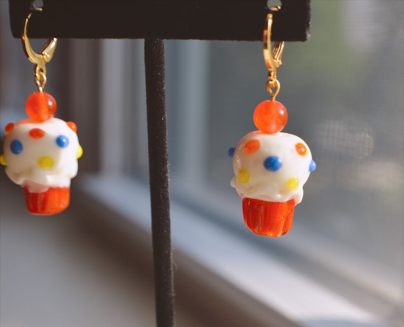 May include: A pair of whimsical cupcake earrings. Each earring features a white frosted cupcake with colorful polka dots, a red cupcake liner, and a small orange bead. The earrings have gold-colored leverback closures.