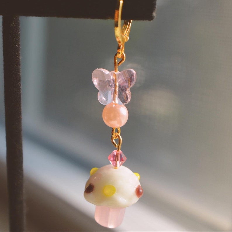 May include: A single gold-colored earring with a whimsical design. It features a pink butterfly, a peach-colored bead, a pink crystal, and a white mushroom charm with brown and yellow accents. The earring has a lever-back closure.
