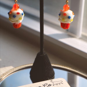May include: A pair of cupcake-shaped earrings with white frosting, colorful sprinkles, and a red cherry on top. The earrings have a red cupcake base and gold-colored ear wires. The earrings are displayed on a black stand with a reflective surface.