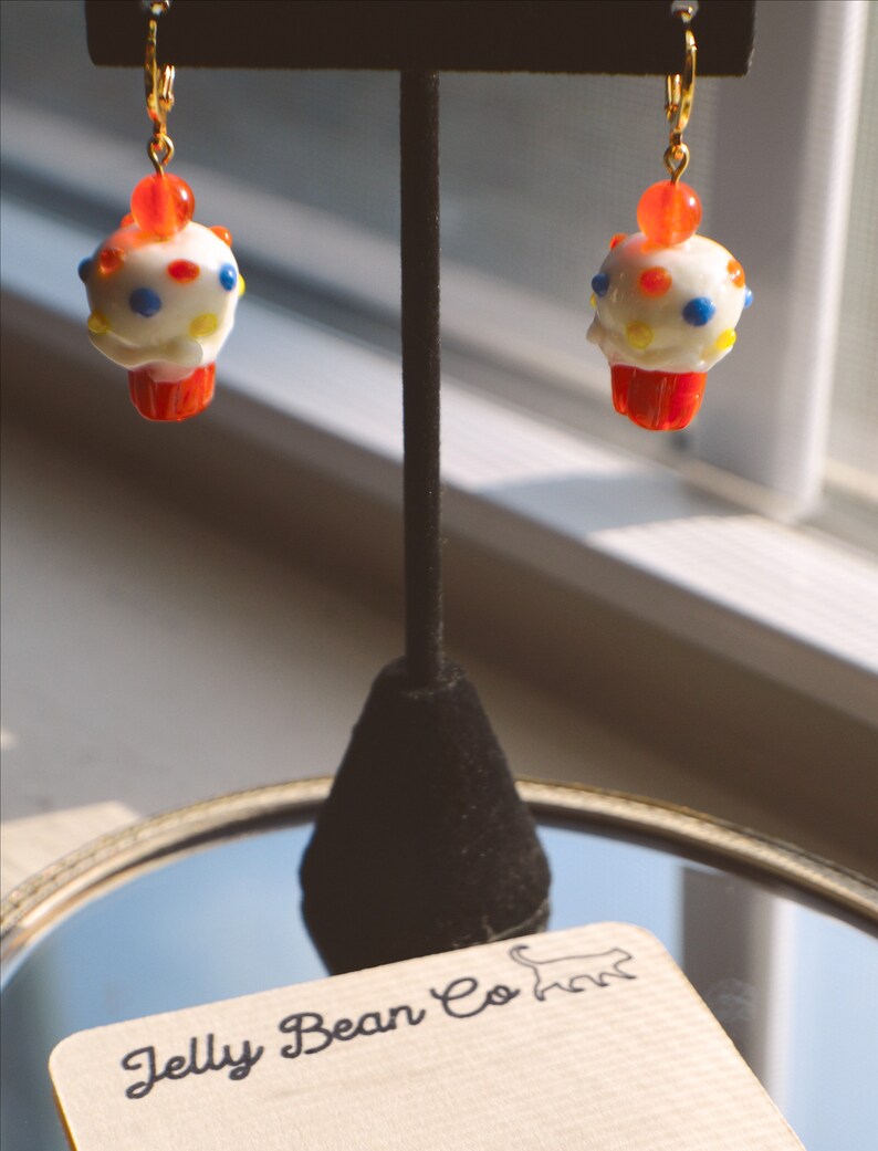 May include: A pair of cupcake-shaped earrings with white frosting, colorful sprinkles, and a red cherry on top. The earrings have a red cupcake base and gold-colored ear wires. The earrings are displayed on a black stand with a reflective surface.