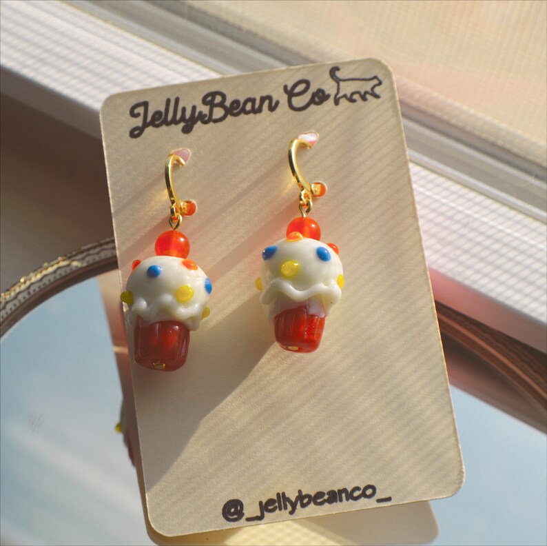 May include: A pair of cupcake-shaped earrings. Each earring features a red base, white frosting with colorful sprinkles, and a red cherry on top. The earrings hang from gold-colored hoops. The earrings are displayed on a tan card with the text "JellyBean Co" and the Instagram handle "@_jellybeanco_".