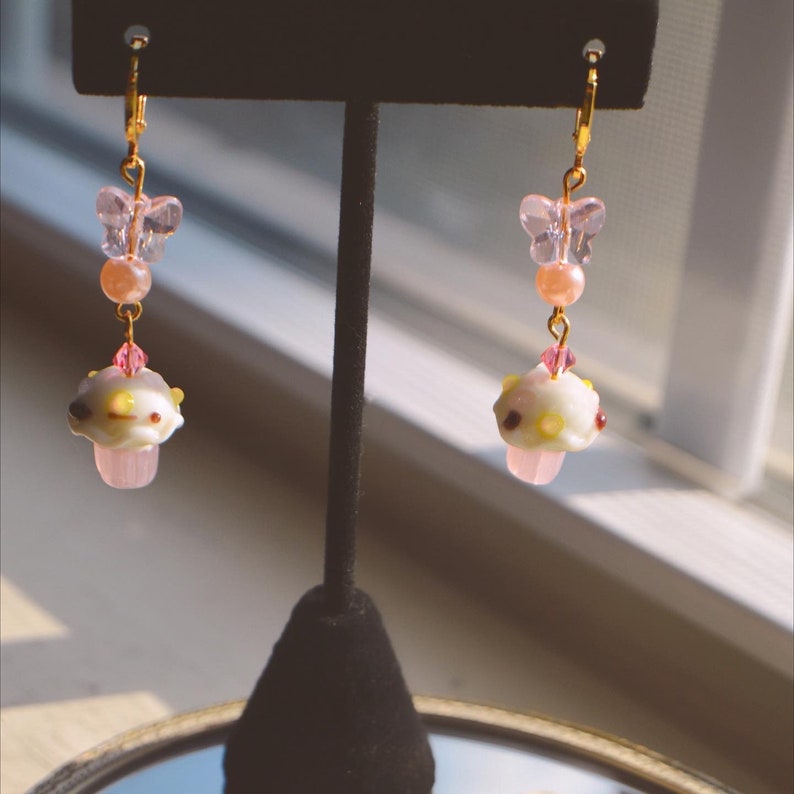 May include: A pair of dangle earrings with a whimsical design. Each earring features a gold-colored leverback, a pink butterfly charm, a peach-colored bead, and a mushroom-shaped charm with white, yellow, and brown details. The mushroom sits atop a pink base.