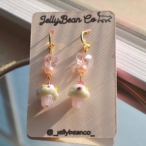 May include: A pair of gold-colored hoop earrings with pink and white mushroom charms. The earrings also feature pink beads, butterfly-shaped charms, and the text "JellyBean Co" on the product tag. The earrings are displayed on a light-colored card.