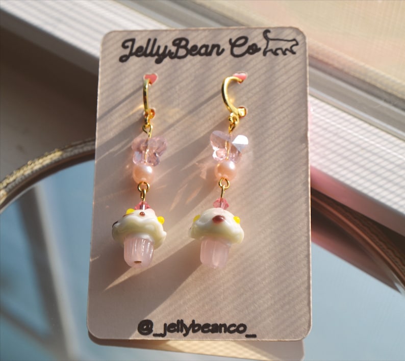 May include: A pair of gold-colored hoop earrings with pink and white mushroom charms. The earrings also feature pink beads, butterfly-shaped charms, and the text "JellyBean Co" on the product tag. The earrings are displayed on a light-colored card.