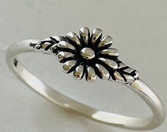 Könnte beinhalten: Ein silberner Ring mit einem zarten Gänseblümchen-Design. Die Gänseblümchen haben eine kleine Mitte und leicht erhabene Blütenblätter. Der Ring ist schlicht und elegant.