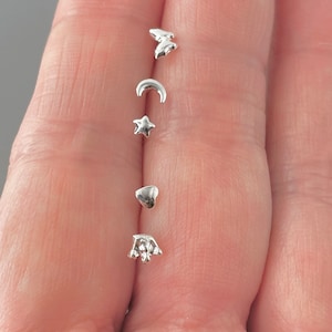 Puede incluir: Cinco pendientes de plata con abalorios, cada uno con un diseño diferente: una mariposa, una luna creciente, una estrella, un corazón y una corona.