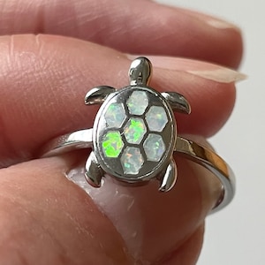 Puede incluir: Un anillo de plata con un dije de tortuga de ópalo blanco. El caparazón de la tortuga está hecho de pequeñas piezas hexagonales de ópalo.