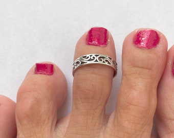 May include: A silver toe ring with a delicate, ornate design. The ring is worn on the second toe of a foot with pink nail polish.