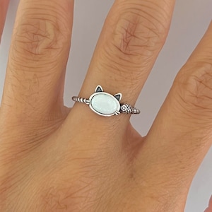 May include: A silver ring featuring a white oval stone set in the shape of a cat's head. The cat is holding a small fish in its mouth.