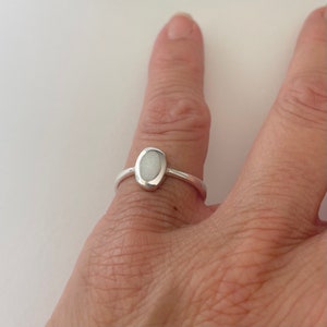 May include: A silver ring with an oval, off-white stone. The ring has a simple band and a bezel setting that holds the stone securely. The ring is shown on a finger against a white background.