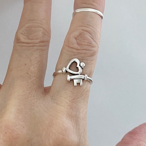 Puede incluir: Un anillo de plata con un diseño de llave en forma de corazón. El anillo se lleva en un dedo.