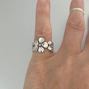 Peut inclure: Une bague en argent avec un motif de plusieurs fleurs. La bague est portée à un doigt.