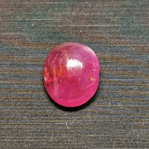 Op de afbeelding: Een ronde, cabochon geslepen robijn met een diepe roze kleur. De steen is geplaatst tegen een getextureerde, metalen achtergrond.