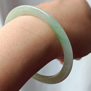 Può includere: Un braccialetto a cerchio di giada verde chiaro. Il braccialetto circolare è indossato su un braccio. La giada ha una qualità traslucida con sottili variazioni di colore, tra cui aree di verde più scuro. Il braccialetto è un gioiello, di circa 6,5 cm di diametro.