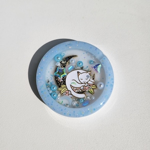 May include: A light blue resin phone grip with a white cat sleeping on a crescent moon. The moon is surrounded by glitter, iridescent rhinestones, and white pearls.