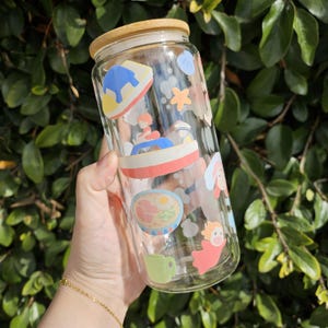 K&ouml;nnte beinhalten: Ein klares Glas mit Bambusdeckel, verziert mit bunten Cartoon-Illustrationen. Der Becher hat eine zylindrische Form und wird vor einem Hintergrund aus gr&uuml;nem Laub gehalten. Die Illustrationen zeigen Charaktere und Essen.
