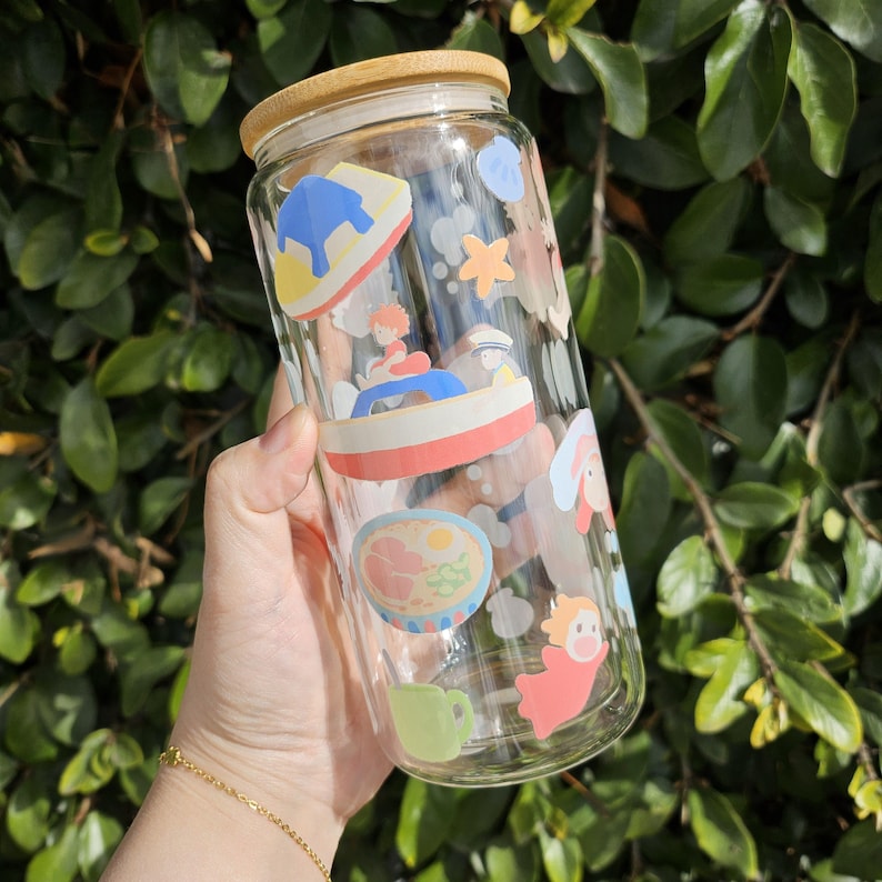K&ouml;nnte beinhalten: Ein klares Glas mit Bambusdeckel, verziert mit bunten Cartoon-Illustrationen. Der Becher hat eine zylindrische Form und wird vor einem Hintergrund aus gr&uuml;nem Laub gehalten. Die Illustrationen zeigen Charaktere und Essen.