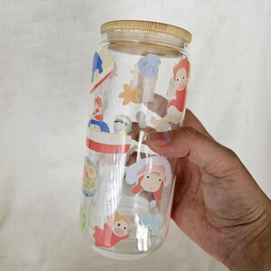 K&ouml;nnte beinhalten: Ein klares Trinkglas mit Bambusdeckel. Das Glas ist mit bunten Cartoon-Charakter-Aufklebern verziert. Das Glas ist zylindrisch und etwa 15 cm hoch.