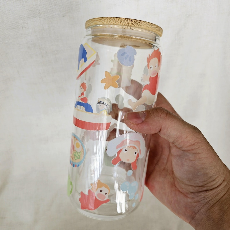 K&ouml;nnte beinhalten: Ein klares Trinkglas mit Bambusdeckel. Das Glas ist mit bunten Cartoon-Charakter-Aufklebern verziert. Das Glas ist zylindrisch und etwa 15 cm hoch.
