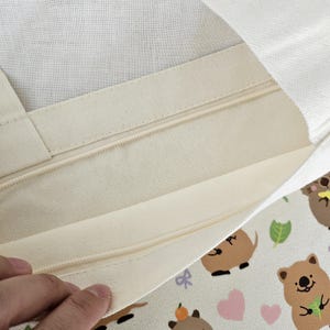 May include: Cream-colored tote bag with a zipper closure. The interior is visible, showing the lining and zipper detail. The exterior features a repeating pattern of cartoon quokkas, hearts, and leaves.