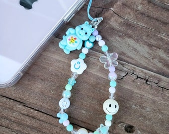 May include: A blue and white beaded phone charm with a smiling face, a star, a butterfly, and a Care Bear character.