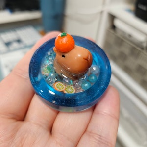 Puede incluir: Una pequeña figura de capibara marrón que lleva un sombrero de fruta naranja, sentada en una piscina de resina azul con cuentas transparentes y azules, y rodajas de naranja.