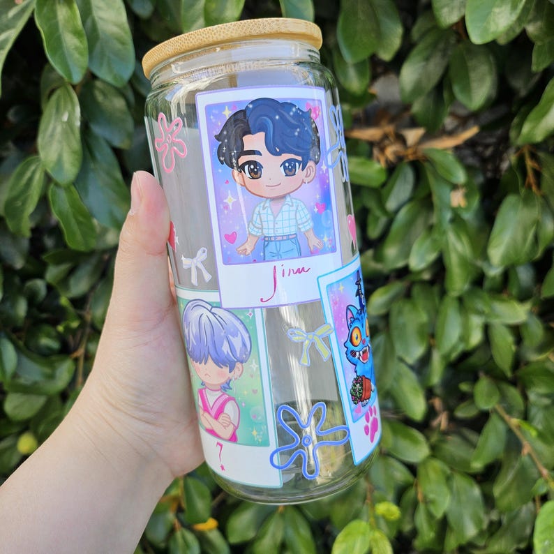 May include: A clear glass tumbler with a bamboo lid, decorated with cartoon images. The tumbler features a cartoon character with blue and black hair, and the name "Jinu" is written below the image. Other cartoon images are also visible.