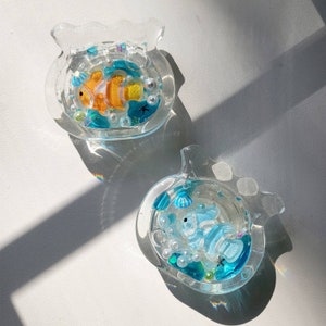 May include: Two clear resin fish bowls with colorful resin fish, seashells, and beads inside. The bowls are shaped like fish and have a white background.