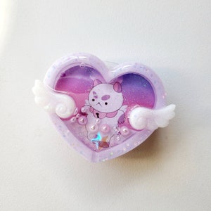 May include: A purple heart-shaped resin charm with a white cat character inside. The cat has a pink nose and a purple star on its chest. The charm is filled with glitter and iridescent beads.