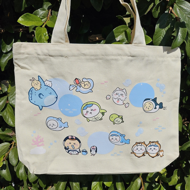 May include: Cream-colored tote bag featuring a whimsical design of cartoon sea creatures and characters in blue, green, and brown. The bag has a simple, rectangular shape with two beige handles, perfect for carrying everyday items.