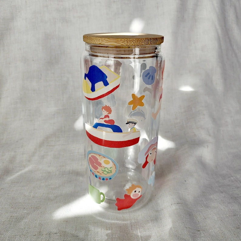 K&ouml;nnte beinhalten: Ein durchsichtiger Glasbecher mit Bambusdeckel, verziert mit bunten Cartoon-Illustrationen. Die Illustrationen umfassen ein Boot, einen Seestern und andere skurrile Designs. Der Becher ist f&uuml;r kalte Getr&auml;nke konzipiert.