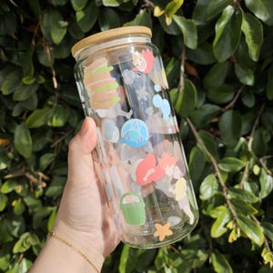 K&ouml;nnte beinhalten: Klarer Glasbecher mit Bambusdeckel, verziert mit bunten Cartoon-Illustrationen. Das Design umfasst Bilder von Charakteren, Essen und anderen Elementen. Der Becher wird vor einem Hintergrund aus gr&uuml;nen Bl&auml;ttern gehalten.