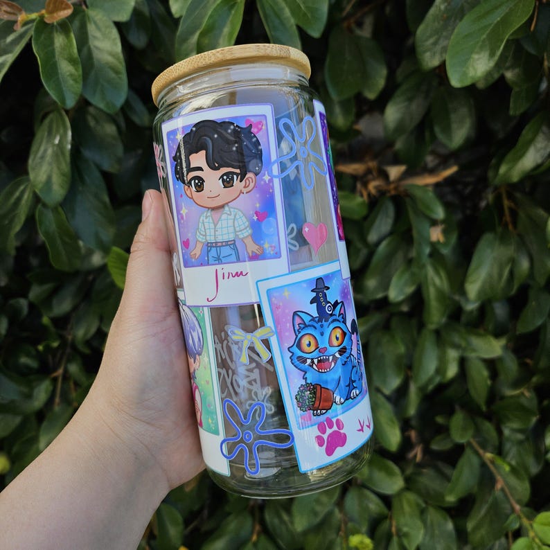 May include: A clear glass tumbler with a bamboo lid, decorated with cartoon images. The tumbler features a cartoon of a person and a blue cat with a hat. The tumbler is held in front of a green leafy background.
