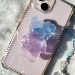 May include: A clear, iridescent purple and blue gummy bear phone charm with glitter. The charm is attached to a clear phone case.