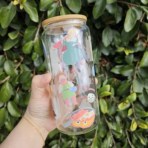 Puede incluir: Una botella de vidrio transparente con tapa de bambú, decorada con pegatinas de dibujos animados de colores. Las pegatinas representan personajes de una película de animación popular, entre ellos una niña con el pelo largo, un niño con un sombrero azul y un personaje con una sartén.