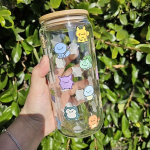 May include: A clear glass beverage can with a bamboo lid, decorated with colorful cartoon character stickers. The stickers are of various shapes and colors. The can is held in front of a green leafy background.