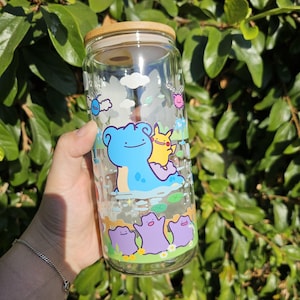 Könnte beinhalten: Ein durchsichtiger Glasbecher mit Bambusdeckel, der bunte Zeichentrickfiguren zeigt. Das Design umfasst eine blaue Kreatur mit einer gelben Figur und lila Kreaturen am Boden. Der Becher ist etwa 15 cm hoch.
