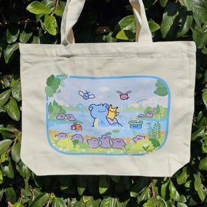 May include: Cream-colored canvas tote bag featuring a colorful illustration of cartoon characters in a serene landscape. The artwork includes a blue lake, a rainbow, and various whimsical creatures. The bag has sturdy handles.