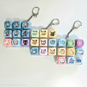 May include: Three square keycap keychains featuring various colorful cartoon characters. Each keychain consists of a grid of nine individual plastic keycaps in pastel and blue tones, each printed with a unique character illustration and attached to a metal ring.